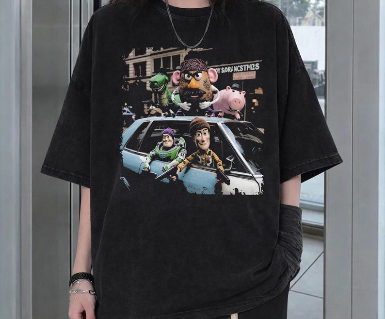 K&ouml;nnte beinhalten: Ein schwarzes T-Shirt mit kurzen &Auml;rmeln und einem Grafikdruck von Toy Story-Figuren. Das Design zeigt eine Gruppe von Charakteren, darunter eine kartoffelf&ouml;rmige Figur, einen gr&uuml;nen Dinosaurier und ein Schwein, die aus einem hellblauen Auto schauen.