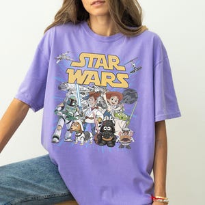 Puede incluir: Camiseta de color lila con un dise&ntilde;o de Star Wars. El dise&ntilde;o presenta el logotipo de Star Wars en amarillo, junto con varios personajes y naves espaciales de la serie de pel&iacute;culas.
