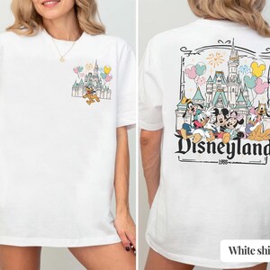 Pode incluir: Camiseta branca com um gr&aacute;fico da Disneyland. A frente mostra Pluto correndo em frente a um castelo com bal&otilde;es. A parte de tr&aacute;s apresenta um gr&aacute;fico maior do castelo da Disneyland com Mickey, Minnie e amigos, com o texto 'Disneyland 1955'.