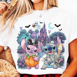 May include: A white t-shirt featuring a graphic print of two blue and pink alien characters in Halloween costumes. One character wears a pumpkin suit, the other is wrapped in bandages. The background shows a castle, park icons, and bats.