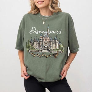 Puede incluir: Camiseta verde salvia con una ilustraci&oacute;n de acuarela de un castillo y el texto "Disneyworld". El dise&ntilde;o incluye patos y patitos. La camiseta tiene mangas cortas y un corte holgado.