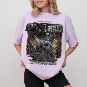 May include: A lilac t-shirt with a graphic from "The Mandalorian" Star Wars series. The design features Grogu and the Mandalorian, with the phrase "THIS IS THE WAY."