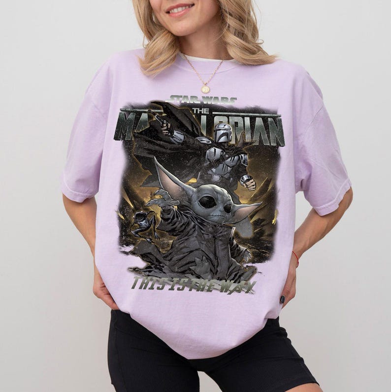 May include: A lilac t-shirt with a graphic from "The Mandalorian" Star Wars series. The design features Grogu and the Mandalorian, with the phrase "THIS IS THE WAY."