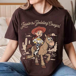May include: A dark brown short-sleeve t-shirt featuring a graphic print of Jessie the Yodeling Cowgirl riding a horse. The design includes desert scenery with cacti and skulls, with the text Jessie the Yodeling Cowgirl printed above.