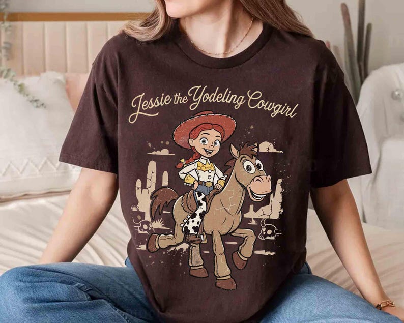 May include: A dark brown short-sleeve t-shirt featuring a graphic print of Jessie the Yodeling Cowgirl riding a horse. The design includes desert scenery with cacti and skulls, with the text Jessie the Yodeling Cowgirl printed above.