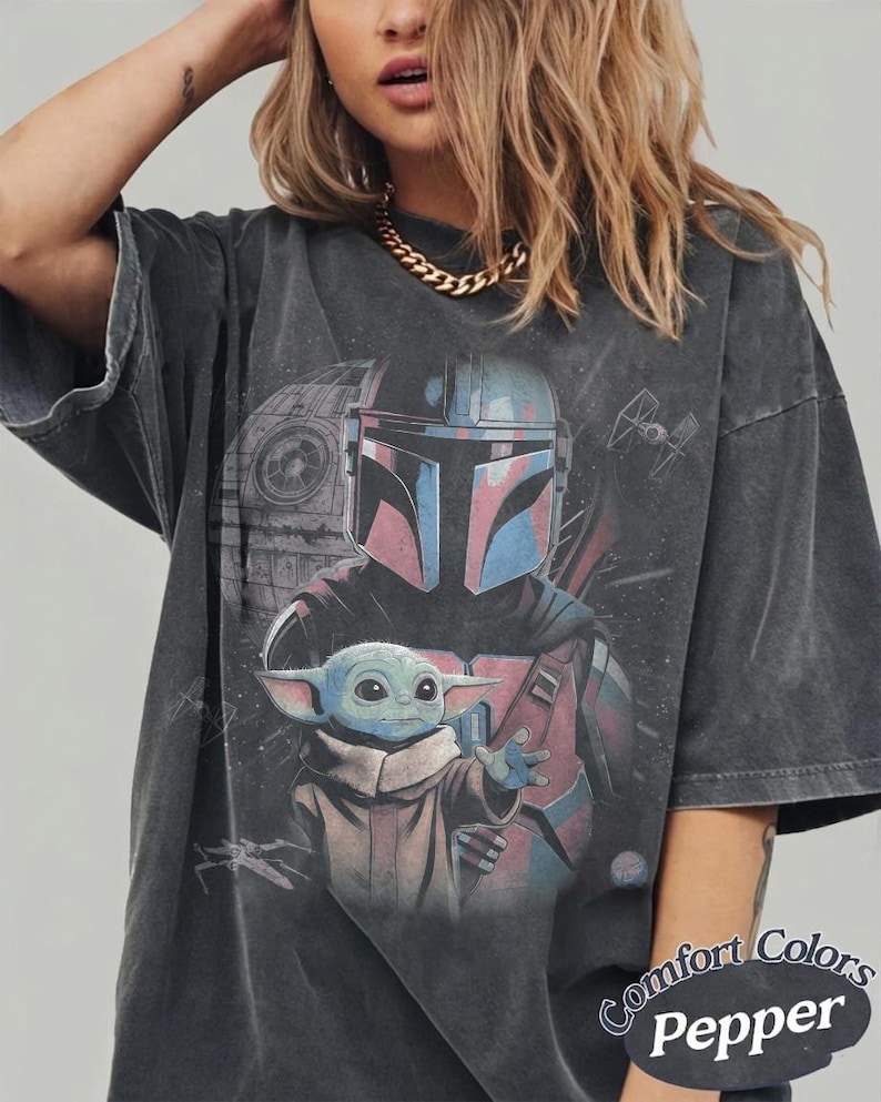 May include: A charcoal gray oversized t-shirt featuring a graphic print of a Mandalorian helmet and a small green creature with large ears. The background includes a space station and starships. The text Comfort Colors Pepper is printed at the bottom.