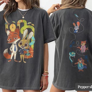 May include: Dark gray t-shirt featuring a colorful graphic of a fox and a rabbit, with the text "Pepper shirt" at the bottom. The back of the shirt has cartoon characters. The shirt is made of a soft material.