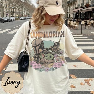 Puede incluir: Camiseta de color marfil con un dise&ntilde;o de The Mandalorian, Grogu y una nave espacial, rodeados de flores. El texto "MANDALORIAN" est&aacute; impreso encima del dise&ntilde;o. La camiseta se lleva con una gorra beige y un bolso negro.