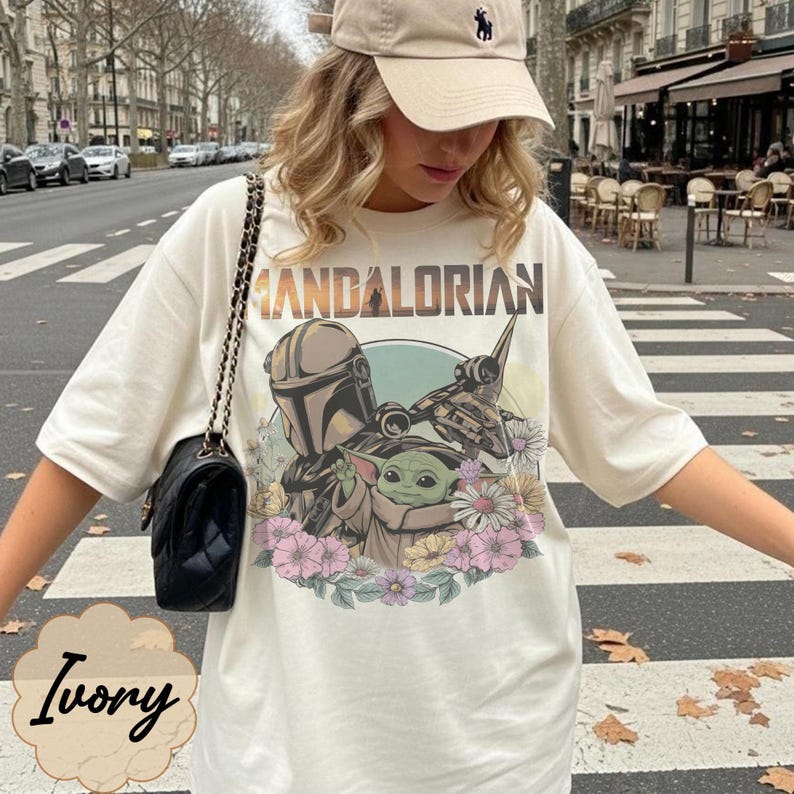 Puede incluir: Camiseta de color marfil con un dise&ntilde;o de The Mandalorian, Grogu y una nave espacial, rodeados de flores. El texto "MANDALORIAN" est&aacute; impreso encima del dise&ntilde;o. La camiseta se lleva con una gorra beige y un bolso negro.