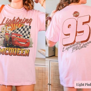 May include: Light pink t-shirt featuring a graphic of Lightning McQueen from the movie Cars. The front design includes the text "Lightning McQueen 95" with a checkered flag and a sunburst background. The back of the shirt displays a large "95" and "Lightning McQueen".