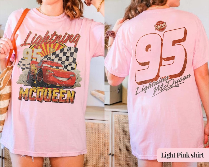 May include: Light pink t-shirt featuring a graphic of Lightning McQueen from the movie Cars. The front design includes the text "Lightning McQueen 95" with a checkered flag and a sunburst background. The back of the shirt displays a large "95" and "Lightning McQueen".