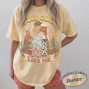 Puede incluir: Una camiseta de manga corta color amarillo mantequilla con un estampado gr&aacute;fico de dos figuras con sombreros de vaquero en un paisaje des&eacute;rtico con cactus y rosas. El texto dice COW GIRL LIKE ME. La camiseta tiene cuello redondo.