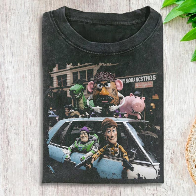 K&ouml;nnte beinhalten: Ein anthrazitfarbenes T-Shirt mit einem Grafikdruck von Charakteren aus der Toy Story-Reihe. Das Design zeigt eine kartoffelf&ouml;rmige Figur, einen gr&uuml;nen Dinosaurier, ein rosa Schwein und zwei Weltraum-Ranger-Figuren in und um ein hellblaues Auto.