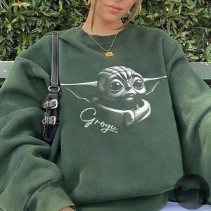 May include: Dark green sweatshirt featuring a detailed graphic of Grogu, also known as Baby Yoda, from The Mandalorian. The image is in grayscale with the name "Grogu" written below in a cursive font. The sweatshirt has a crew neck and long sleeves.
