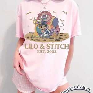 May include: A light pink t-shirt featuring a graphic of characters from Lilo & Stitch, with the text "LILO & STITCH EST. 2002". The shirt is a Comfort Colors Blossom tee.