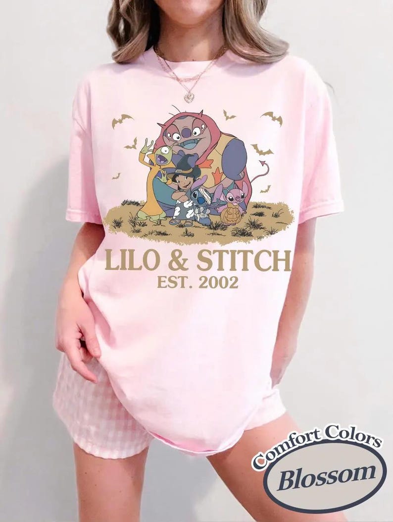 May include: A light pink t-shirt featuring a graphic of characters from Lilo & Stitch, with the text "LILO & STITCH EST. 2002". The shirt is a Comfort Colors Blossom tee.