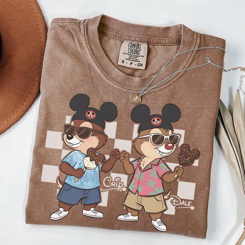 Pu&ograve; includere: T-shirt marrone con i personaggi dei cartoni animati Cip e Ciop con occhiali da sole e gelati. La maglietta ha uno sfondo a scacchi e le parole "Chip" e "Dale" sono stampate.