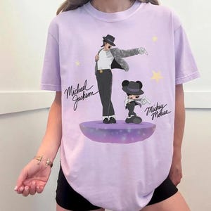 Puede incluir: Una camiseta de color lila claro con un gr&aacute;fico de Michael Jackson y Mickey Mouse. La ilustraci&oacute;n muestra a Michael Jackson con sombrero y traje negro, y a Mickey Mouse con un atuendo similar. Las palabras "Michael Jackson" y "Mickey Mouse" est&aacute;n escritas en cursiva.
