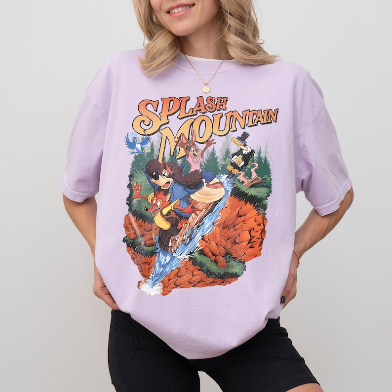May include: A lilac t-shirt with a vibrant graphic of Splash Mountain characters. The design includes the text "SPLASH MOUNTAIN" in orange and yellow, with cartoon figures on a log flume. The shirt has a relaxed, oversized style.