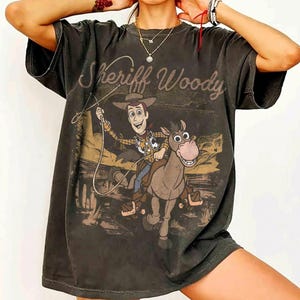 K&ouml;nnte beinhalten: Dunkelgraues T-Shirt mit einem verblichenen Motiv von Sheriff Woody aus Toy Story auf einem Pferd und dem Text "Sheriff Woody". Das T-Shirt hat kurze &Auml;rmel und eine lockere Passform mit Vintage-&Auml;sthetik. Das Design ist auf der Vorderseite aufgedruckt.