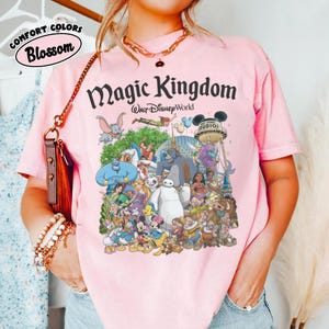 Puede incluir: Camiseta rosa claro con un estampado que presenta varios personajes de Disney y el texto "Magic Kingdom Walt Disney World". La camiseta tambi&eacute;n tiene el texto "Comfort Colors Blossom".