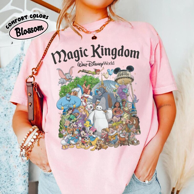 Puede incluir: Camiseta rosa claro con un estampado que presenta varios personajes de Disney y el texto "Magic Kingdom Walt Disney World". La camiseta tambi&eacute;n tiene el texto "Comfort Colors Blossom".