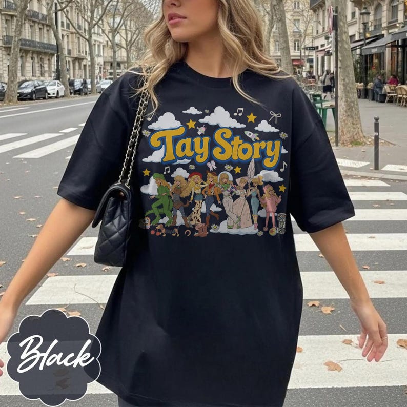 May include: A black short-sleeved t-shirt featuring a graphic print with the text Tay Story in yellow letters. The design includes a row of illustrated characters, white clouds, yellow stars, musical notes, and a rocket ship on a black background.