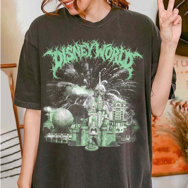 K&ouml;nnte beinhalten: Dunkelgraues T-Shirt mit einem gr&uuml;nen Aufdruck "DISNEY WORLD" in einer stacheligen Schriftart. Die Grafik zeigt ein Schloss, Feuerwerk und einen Golfball. Das Shirt hat eine lockere Passform.