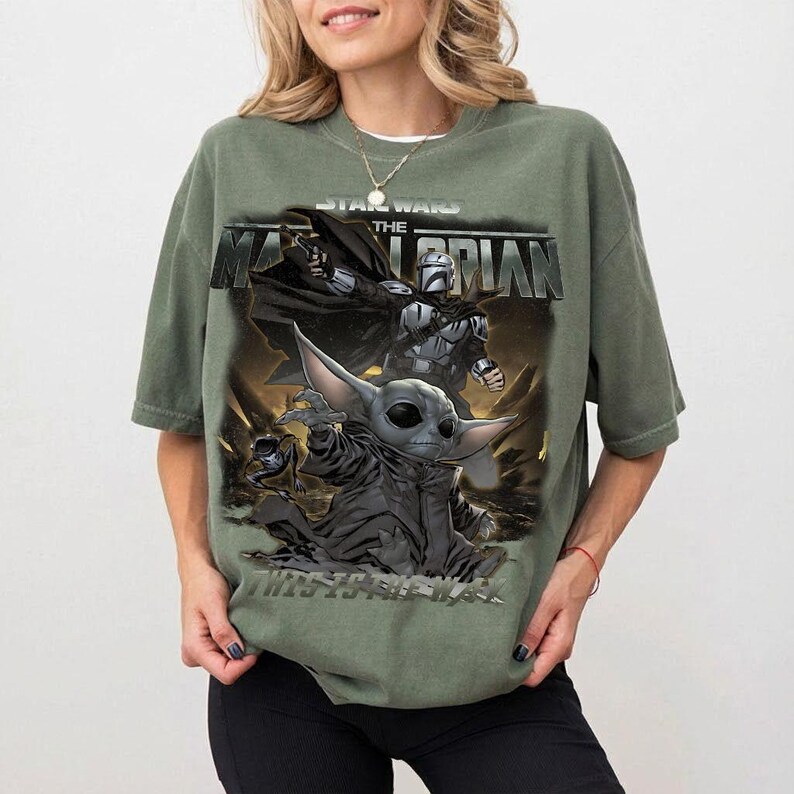 May include: Olive green t-shirt with a graphic from "The Mandalorian" Star Wars series. The design features the Mandalorian, Grogu, and the show's title. The shirt has short sleeves and a relaxed fit.