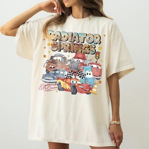 May include: Cream-colored t-shirt featuring a colorful graphic of characters from the movie Cars, including Lightning McQueen. The design includes the text "Radiator Springs" and "Lightning McQueen". The shirt has short sleeves and a relaxed fit.