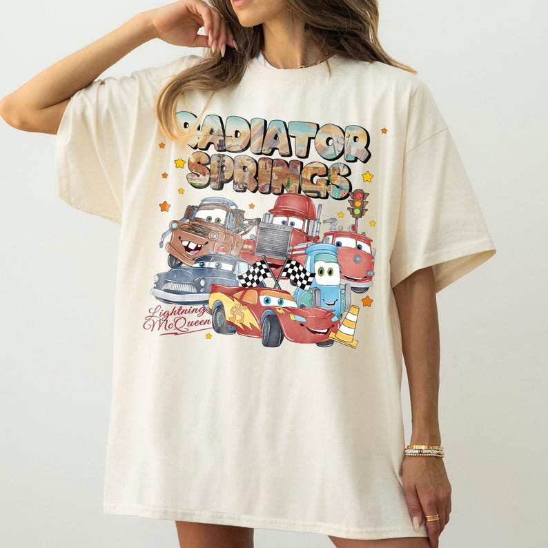 May include: Cream-colored t-shirt featuring a colorful graphic of characters from the movie Cars, including Lightning McQueen. The design includes the text "Radiator Springs" and "Lightning McQueen". The shirt has short sleeves and a relaxed fit.
