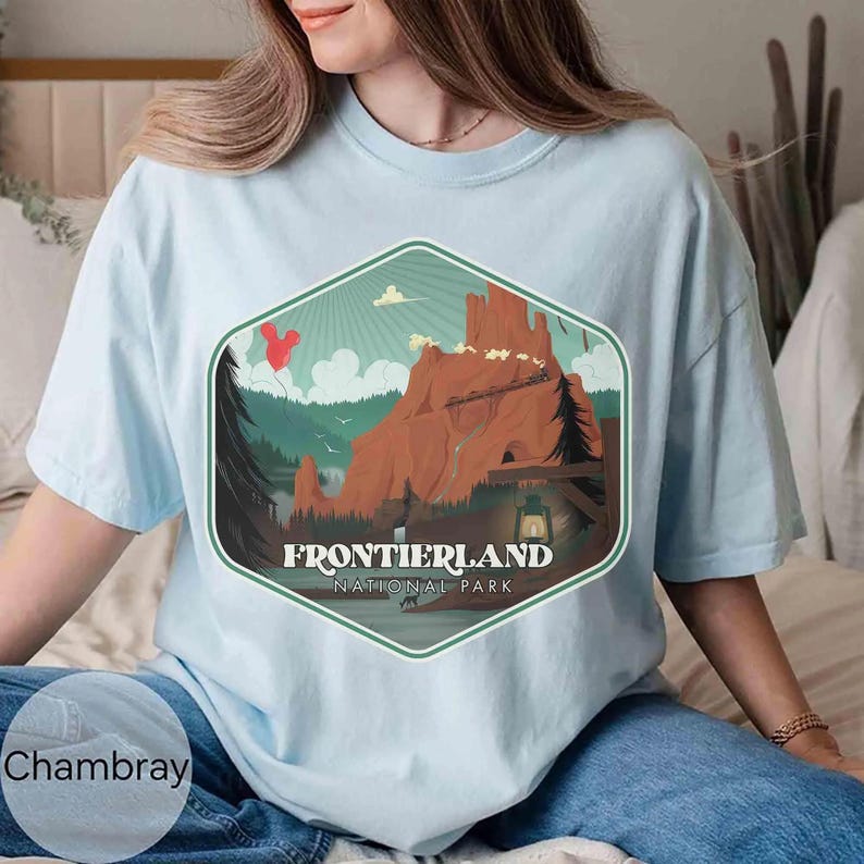 Op de afbeelding: Een lichtblauw t-shirt met een zeshoekige grafische print van een berglandschap. Het ontwerp bevat een rode ballon, een trein op een spoor, een lantaarn en de tekst "FRONTIERLAND NATIONAL PARK" in witte, vetgedrukte, schreefloze letters.