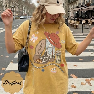Puede incluir: Una camiseta de color amarillo mostaza con un estampado gr&aacute;fico de una bota vaquera con manchas de vaca, un sombrero rojo y bloques con las letras T, S y el n&uacute;mero 13. El dise&ntilde;o incluye el nombre Taylor, un lazo, flores y el texto I KNEW IT WHEN YOU.