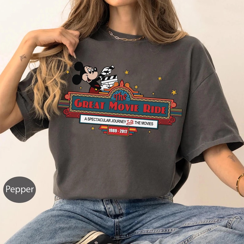 Puede incluir: Camiseta gris oscuro con Mickey Mouse sosteniendo una claqueta de cine. La camiseta muestra el texto "The Great Movie Ride" en un dise&ntilde;o de marquesina, con el texto: "A Spectacular Journey Into The Movies 1989-2017."