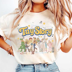 May include: A cream-colored t-shirt featuring the text Tay Story in large yellow letters with a blue outline. Below the text is a graphic illustration of several blonde figures in various outfits, surrounded by small stars, musical notes, and clouds.