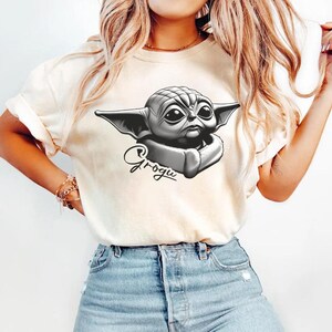 May include: Cream-colored t-shirt featuring a black and white illustration of Grogu, with the name Grogu written in cursive below the image. The t-shirt is paired with blue denim jeans.