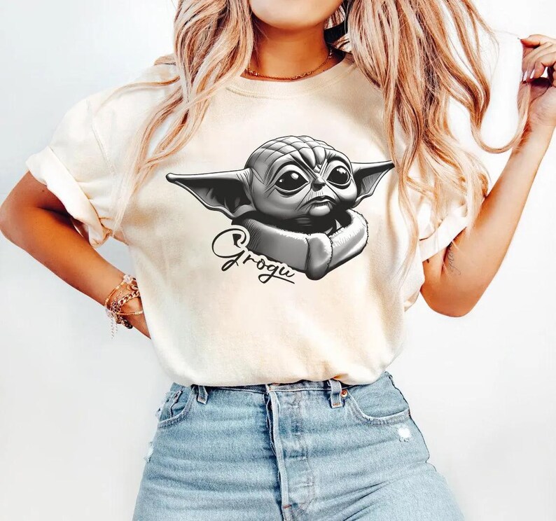 May include: Cream-colored t-shirt featuring a black and white illustration of Grogu, with the name Grogu written in cursive below the image. The t-shirt is paired with blue denim jeans.