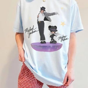 Puede incluir: Camiseta azul claro con un gr&aacute;fico de Michael Jackson y Mickey Mouse. La ilustraci&oacute;n muestra a Michael Jackson con sombrero y traje negro, y a Mickey Mouse con un atuendo similar. La camiseta tiene el texto "Michael Jackson" y "Mickey Mouse".