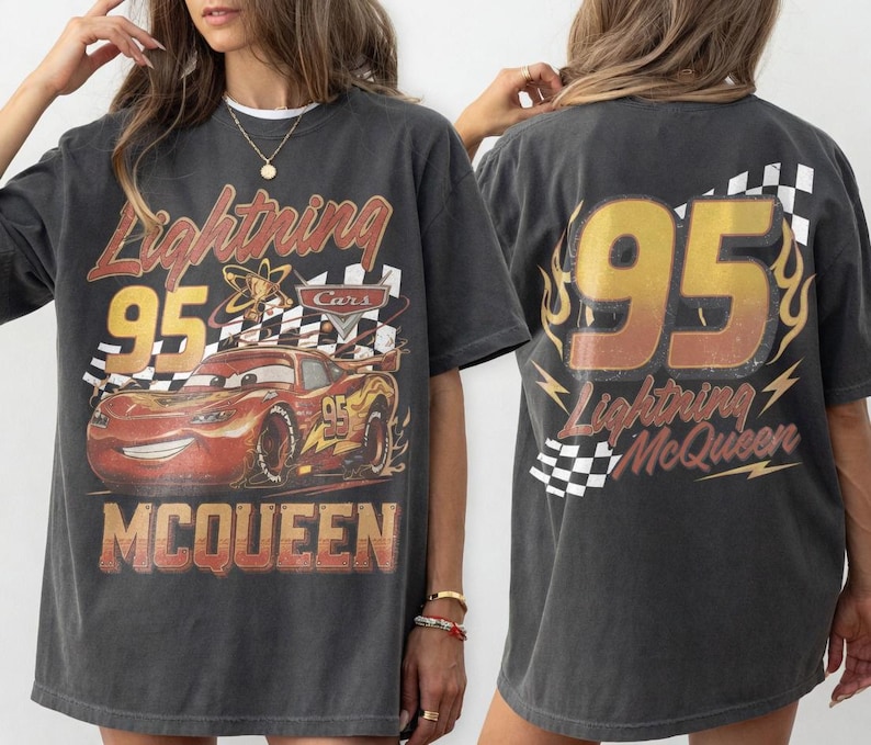 May include: Dark gray t-shirt featuring a graphic of Lightning McQueen from the Cars movie. The front design includes the number 95, the text "Lightning McQueen", and a checkered flag. The back also features the number 95.