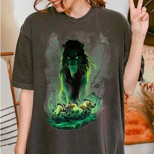 Puede incluir: Camiseta gris oscuro con una ilustraci&oacute;n verde y negra de un le&oacute;n y tres hienas. El le&oacute;n tiene una expresi&oacute;n amenazante, y las hienas parecen estar corriendo. El dise&ntilde;o tiene un efecto de brillo verde y ahumado.