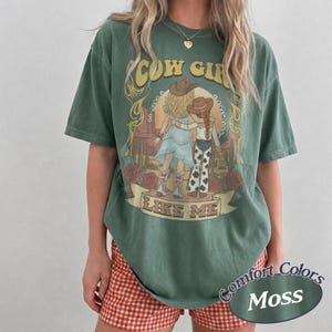 Puede incluir: Una camiseta de manga corta color verde musgo con un estampado gr&aacute;fico de dos figuras con sombreros de vaquero y botas en un paisaje des&eacute;rtico. El texto COW GIRL LIKE ME est&aacute; impreso en amarillo y blanco. Tiene cuello redondo.