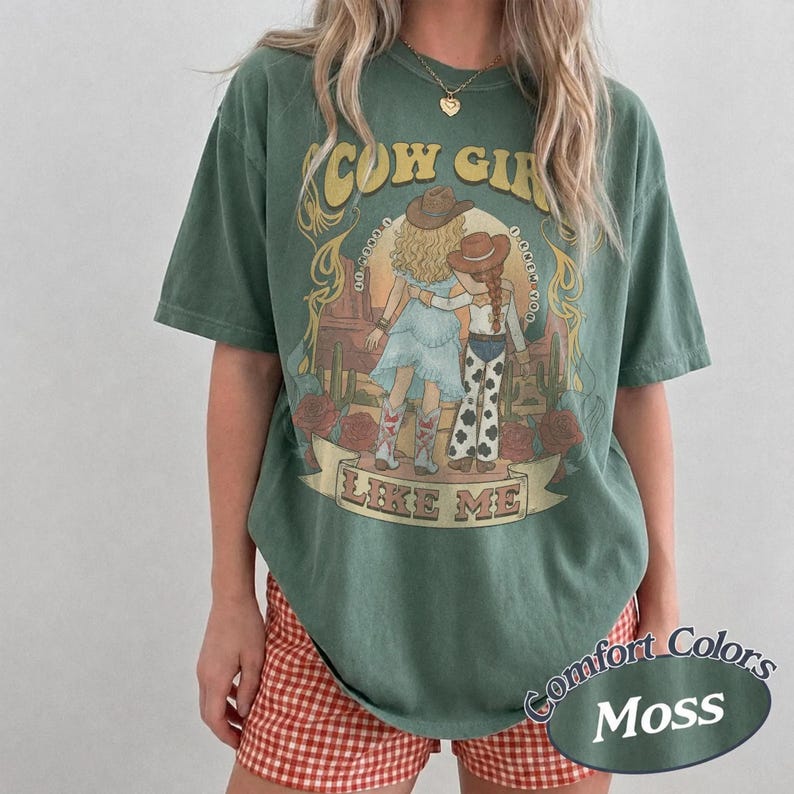Puede incluir: Una camiseta de manga corta color verde musgo con un estampado gr&aacute;fico de dos figuras con sombreros de vaquero y botas en un paisaje des&eacute;rtico. El texto COW GIRL LIKE ME est&aacute; impreso en amarillo y blanco. Tiene cuello redondo.