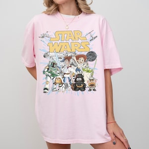 Puede incluir: Camiseta rosa claro con un estampado de personajes de Star Wars y el logo de Star Wars en amarillo. El dise&ntilde;o incluye a Darth Vader, Woody y Buzz Lightyear. La camiseta tiene mangas cortas y un corte holgado.
