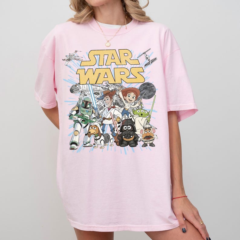 Puede incluir: Camiseta rosa claro con un estampado de personajes de Star Wars y el logo de Star Wars en amarillo. El dise&ntilde;o incluye a Darth Vader, Woody y Buzz Lightyear. La camiseta tiene mangas cortas y un corte holgado.