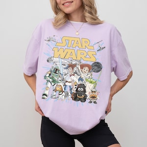 Puede incluir: Una camiseta de color lila claro con un gr&aacute;fico de personajes de Star Wars, incluyendo a Boba Fett, Woody y Buzz Lightyear. Las palabras "STAR WARS" est&aacute;n en letras amarillas sobre los personajes. La camiseta es de corte holgado.