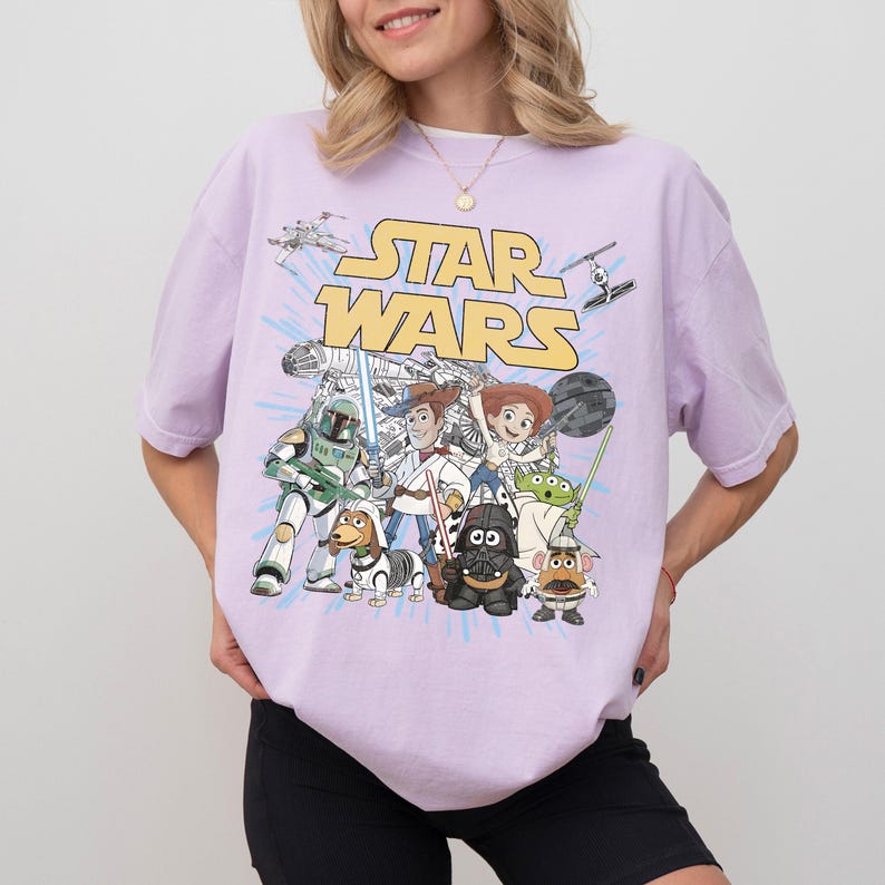 Puede incluir: Una camiseta de color lila claro con un gr&aacute;fico de personajes de Star Wars, incluyendo a Boba Fett, Woody y Buzz Lightyear. Las palabras "STAR WARS" est&aacute;n en letras amarillas sobre los personajes. La camiseta es de corte holgado.