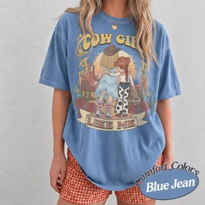 Puede incluir: Una camiseta azul con efecto denim que presenta un estampado gr&aacute;fico de dos figuras con sombreros de vaquero en un paisaje des&eacute;rtico. El texto "COW GIRL" aparece en la parte superior y "LIKE ME" en una cinta en la parte inferior.