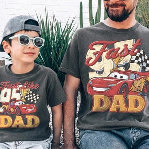 May include: Two matching charcoal gray t-shirts with a vintage-style graphic of a red race car, a trophy, and the words "Fast Like Dad" and "Dad". One shirt is on a child, the other on an adult.