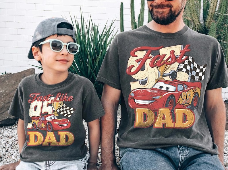 May include: Two matching charcoal gray t-shirts with a vintage-style graphic of a red race car, a trophy, and the words "Fast Like Dad" and "Dad". One shirt is on a child, the other on an adult.