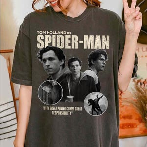 May include: Dark gray t-shirt featuring Tom Holland as Spider-Man. The design includes multiple images of Holland, the Spider-Man logo, and the quote "With great power comes great responsibility."